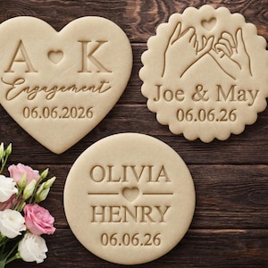 Peut inclure: Trois biscuits beiges avec des motifs en relief sur une surface en bois foncé. Un biscuit en forme de cœur indique "A K Engagement 06.06.2026". Un autre biscuit rond dit "Olivia Henry 06.06.26". Le troisième biscuit montre des mains jointes et dit "Joe & May 06.06.26."