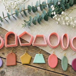 Peut inclure: Ensemble de découpoirs à boucles d'oreilles en pâte polymère rose de différentes formes, comme goutte d'eau, arche et croissant, avec des boucles d'oreilles finies assorties. Présenté sur une surface en bois avec de l'eucalyptus et des fleurs blanches.