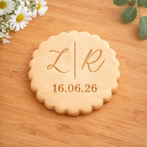 May include: A round, light-brown cookie with a scalloped edge. The cookie is engraved with the letters "Z" and "R" separated by a vertical line, and the date "16.06.26" below. The cookie rests on a wooden surface, with flowers in the background.