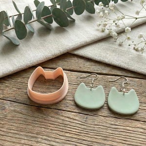 May include: A peach-coloured cat-shaped cookie cutter and two mint green cat-shaped earrings with silver hooks are displayed on a wooden surface. Eucalyptus leaves and white flowers are arranged around the items.