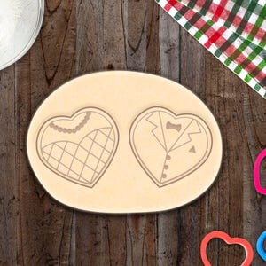 May include: A close-up of a heart-shaped cookie cutter with a tuxedo design on the left and a heart-shaped cookie cutter with a dress design on the right. The cookie cutters are on a wooden surface with a white tablecloth and a bowl of flour in the background.