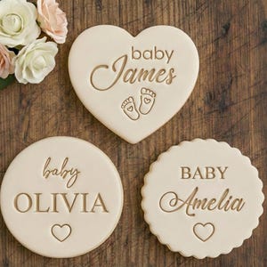 Puede incluir: Tres galletas decorativas para anunciar un bebé. Una tiene forma de corazón con "baby James" y huellas de pies. Otra es redonda con "baby Olivia" y un corazón. La tercera es festoneada con "BABY Amelia" y un corazón.