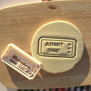 May include: A white plastic cookie cutter in the shape of a movie ticket with the text "ADMIT ONE" and three stars. The cookie cutter is on a wooden cutting board next to a round piece of cookie dough with the same design imprinted on it.
