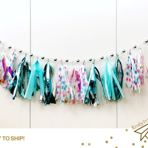 May include: A teal, pink, and white tassel garland with silver beads. The garland is made of tissue paper and is perfect for a party or celebration.