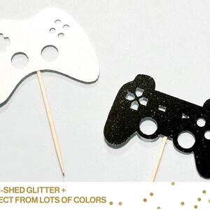 May include: Two black and white glitter game controller cake toppers. The black controller has a sparkly finish. Both toppers are attached to wooden sticks.