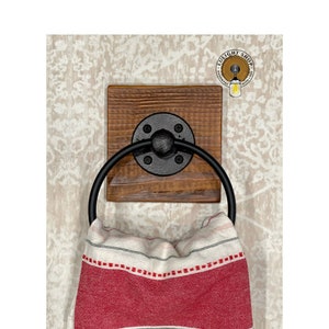 May include: A rustic wooden towel ring with a black metal ring and a black metal flange. The ring is mounted on a wooden board with a natural wood finish. A red and white striped towel is hanging on the ring.