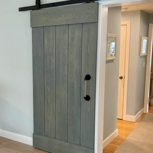 May include: A gray wooden sliding barn door with a rope handle and black hardware. The door is open and reveals a hallway with a white door and a rug on the floor.