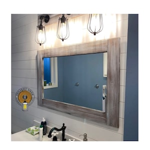 May include: A rectangular wall mirror featuring a weathered, light brown wood frame with a distressed finish. The mirror is mounted on a white horizontal shiplap wall beneath three industrial-style pendant lights with black wire cages.