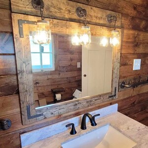 May include: A rectangular bathroom mirror framed in weathered wood with dark metal corner brackets. Three mason jar light fixtures are mounted to the top of the frame. A white marble countertop with a black faucet is positioned below the mirror.