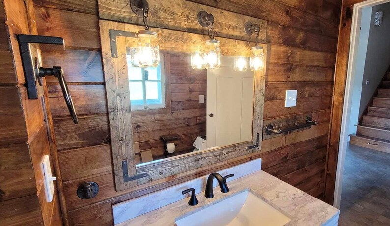 May include: A rectangular bathroom mirror framed in weathered wood with dark metal corner brackets. Three mason jar light fixtures are mounted to the top of the frame. A white marble countertop with a black faucet is positioned below the mirror.