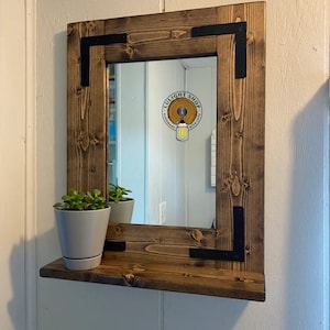 May include: A rustic, square wooden mirror with a shelf, featuring a natural wood grain finish. Black metal corner brackets add a touch of industrial style. A small potted plant sits on the shelf. The mirror reflects the room.