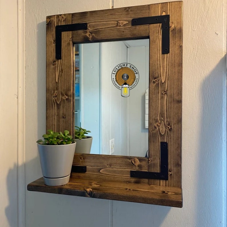 May include: A rustic, square wooden mirror with a shelf, featuring a natural wood grain finish. Black metal corner brackets add a touch of industrial style. A small potted plant sits on the shelf. The mirror reflects the room.