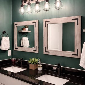May include: A bathroom vanity with a black granite countertop and two white porcelain sinks. The vanity has white cabinets with silver hardware. The bathroom has a dark green wall with two framed mirrors and a light fixture with five caged bulbs. The light fixture is made of black metal pipes and has a rustic industrial style. The text "LULIGHT SHOP HANDMADE WITH LOVE" is visible on the wall.