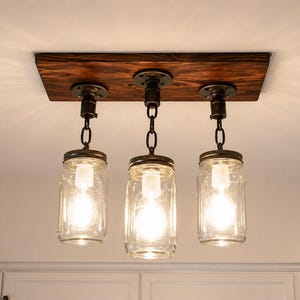 Farmhouse mason jar pendant light fixture with wood beam and chains hanging above rustic dining table in cozy kitchen