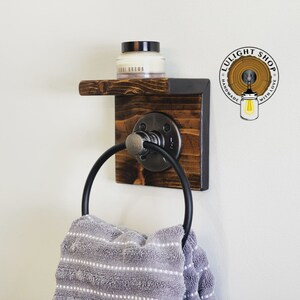 May include: A rustic wooden towel ring with a black metal ring and a small shelf. The shelf has a small jar of cream on it. The towel ring is mounted on a white wall. The text "LULIGHT SHOP HANDMADE WITH LOVE" is visible in the upper right corner of the image.