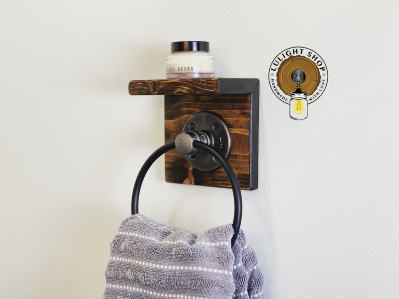 May include: A rustic wooden towel ring with a black metal ring and a small shelf. The shelf has a small jar of cream on it. The towel ring is mounted on a white wall. The text "LULIGHT SHOP HANDMADE WITH LOVE" is visible in the upper right corner of the image.