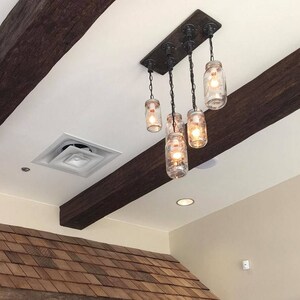 May include: A rustic chandelier featuring five clear glass mason jar lights suspended from a dark wooden beam. The fixture is attached to a rectangular metal plate and hangs from chains. The jars contain warm, glowing light bulbs.