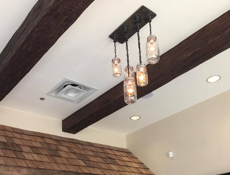 May include: A rustic chandelier featuring five clear glass mason jar lights suspended from a dark wooden beam. The fixture is attached to a rectangular metal plate and hangs from chains. The jars contain warm, glowing light bulbs.