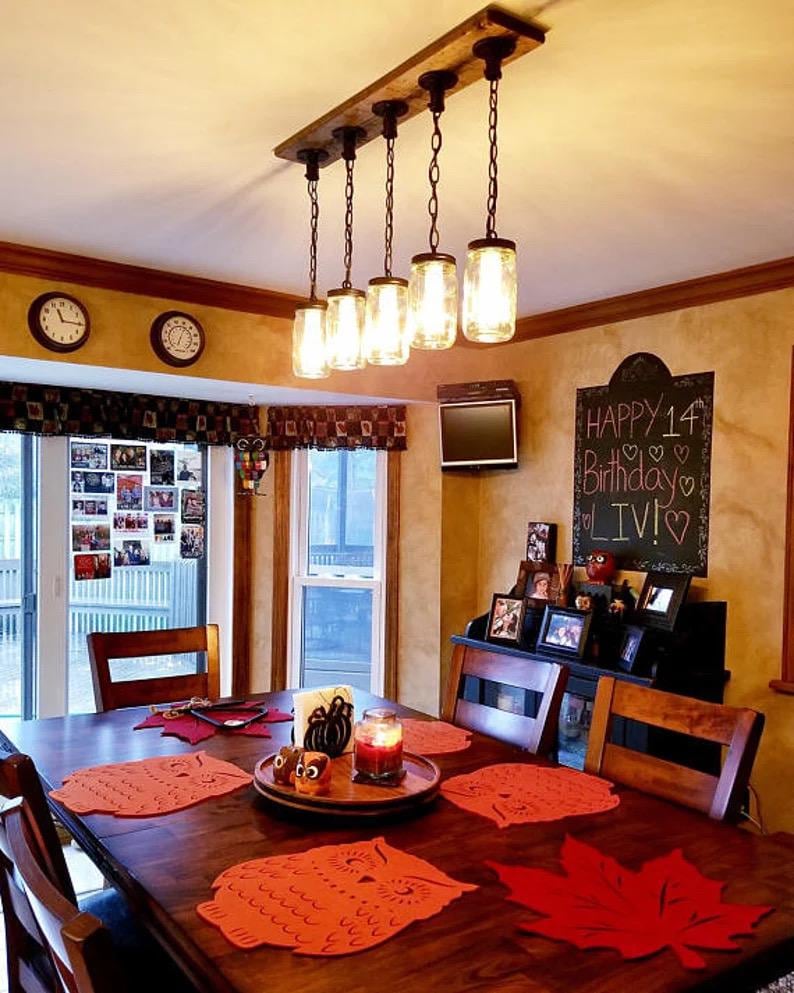 May include: A rustic-style light fixture with three clear glass jar pendant lights suspended from a dark wooden beam. The fixture is mounted above a bathroom vanity with a mirror and sink.