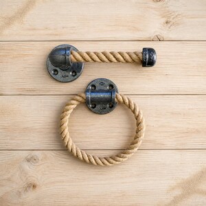 May include: A rustic bathroom set featuring a towel bar and a towel ring. Both pieces have a dark gray metal base and are accented with thick, natural-colored rope. The set is designed to be mounted on a wall.