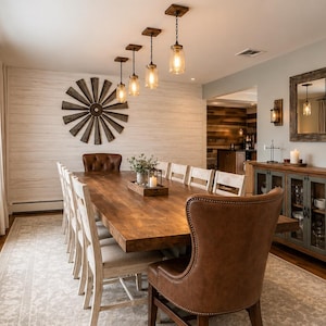May include: A rustic dining room with a long, wooden table set for a large gathering. The room features a windmill wall decor, pendant lights, and a buffet with glass-front cabinets. The chairs are a mix of white and brown leather.