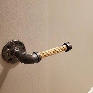 May include: Industrial-style toilet paper holder made from black metal pipes and fittings, featuring a natural rope roll. The holder is mounted on a light-colored wall, showcasing a rustic aesthetic.