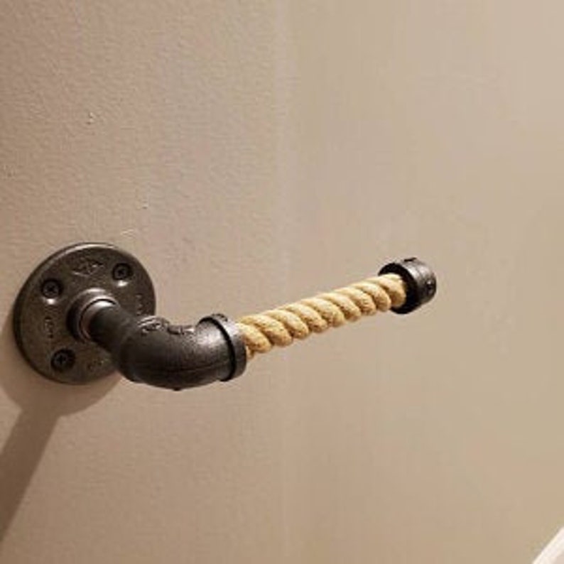 May include: Industrial-style toilet paper holder made from black metal pipes and fittings, featuring a natural rope roll. The holder is mounted on a light-colored wall, showcasing a rustic aesthetic.