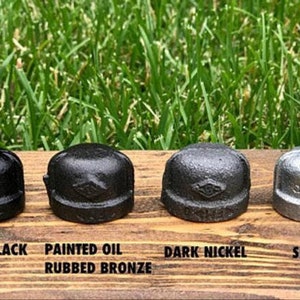 May include: Four metal pipe caps arranged in a row, each featuring a distinct finish: painted black, painted oil rubbed bronze, dark nickel, and silver. Each cap displays a small diamond-shaped logo on the front surface, showcasing industrial hardware.