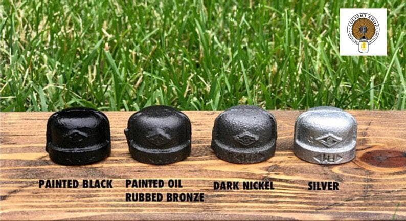 May include: Four metal pipe caps arranged in a row, each featuring a distinct finish: painted black, painted oil rubbed bronze, dark nickel, and silver. Each cap displays a small diamond-shaped logo on the front surface, showcasing industrial hardware.