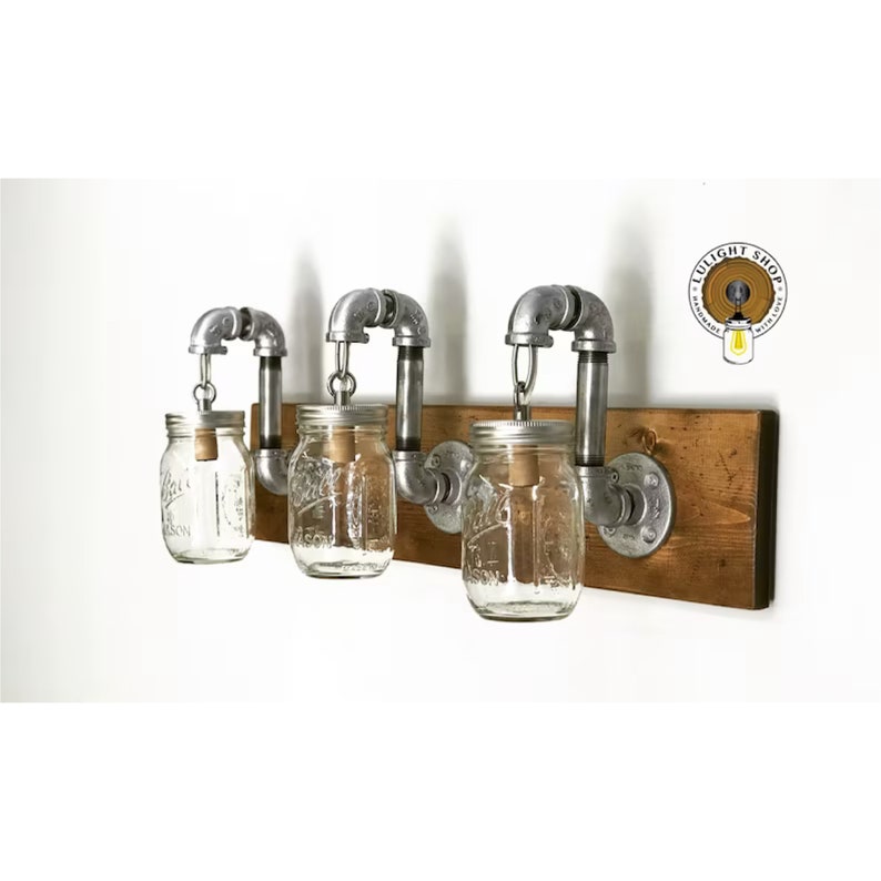 A mason jar light fixture with three clear glass jars hanging from a rustic distressed wood board. Each jar contains a light bulb emitting a warm and cozy glow. The rustic and charming design adds character to any space.