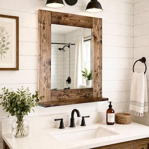 May include: A rustic bathroom vanity with a wooden framed mirror, a white countertop, and dark fixtures. The mirror reflects a shower and window. A framed botanical print and a vase of greenery add decorative touches.