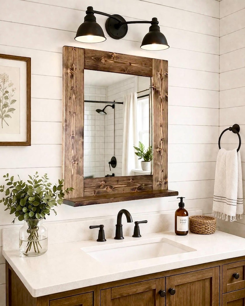 May include: A rustic bathroom vanity with a wooden framed mirror, a white countertop, and dark fixtures. The mirror reflects a shower and window. A framed botanical print and a vase of greenery add decorative touches.