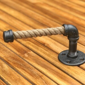 May include: A rustic toilet paper holder made from black iron pipes and a thick, natural rope. The holder has a round base and a right-angle pipe fitting. The rope is a light brown color.