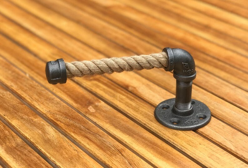 May include: A rustic toilet paper holder made from black iron pipes and a thick, natural rope. The holder has a round base and a right-angle pipe fitting. The rope is a light brown color.