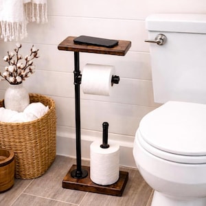 May include: A wooden and metal toilet paper holder with a shelf for a phone. The holder has a dark brown wooden base and shelf, with black metal pipes. It holds a roll of toilet paper and a spare roll. The bathroom decor is modern and functional.