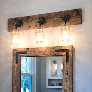 May include: Rustic wooden vanity light fixture with three mason jar lights. The fixture is mounted on a weathered wooden plank. The mirror frame is made of matching wood with black metal corner brackets.