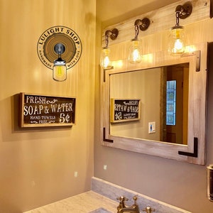 Pu&ograve; includere: Un set specchio e luci per bagno rustico. Il set comprende uno specchio rettangolare con cornice in legno chiaro e tre luci a barattolo Mason. &Egrave; visibile anche un cartello con la scritta "Fresh Soap & Water".