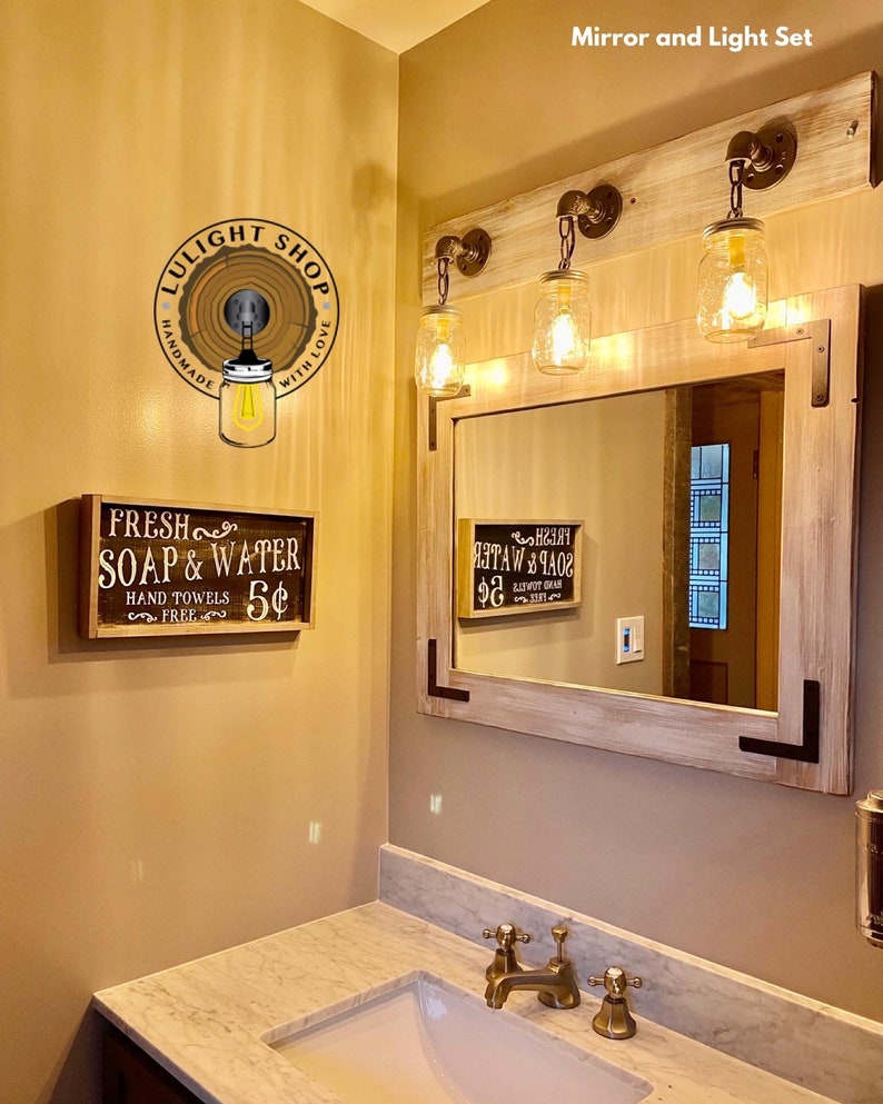 Pu&ograve; includere: Un set specchio e luci per bagno rustico. Il set comprende uno specchio rettangolare con cornice in legno chiaro e tre luci a barattolo Mason. &Egrave; visibile anche un cartello con la scritta "Fresh Soap & Water".