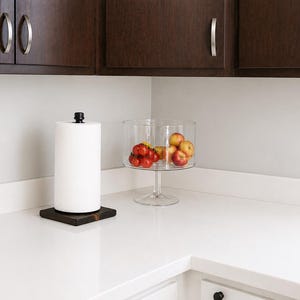 May include: A black metal pipe paper towel holder with a wooden base sits on a white countertop next to a clear glass bowl filled with red tomatoes and yellow apples.
