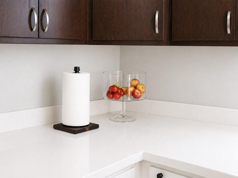 May include: A black metal pipe paper towel holder with a wooden base sits on a white countertop next to a clear glass bowl filled with red tomatoes and yellow apples.