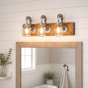 A mason jar light fixture with three clear glass jars hanging from a rustic distressed wood board. Each jar contains a light bulb emitting a warm and cozy glow. The rustic and charming design adds character to any space.