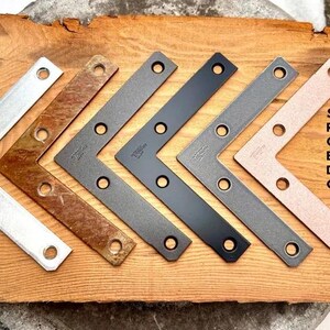 May include: Six metal corner brackets in different finishes: black, oil rubbed bronze, silver, dark nickel, and rusty.  The brackets are arranged in a row from left to right.