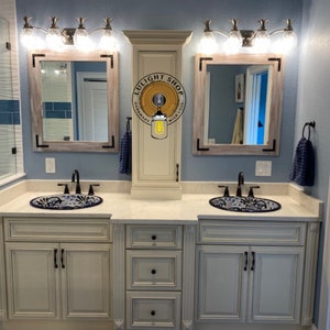 Pu&ograve; includere: Un mobile da bagno con due lavandini, specchi e lampade. Il mobile ha un mobile color crema con ferramenta nera. I lavandini hanno un motivo floreale blu e bianco. Gli specchi hanno una cornice in legno chiaro con accenti angolari neri. Le lampade hanno paralumi in vetro trasparente.
