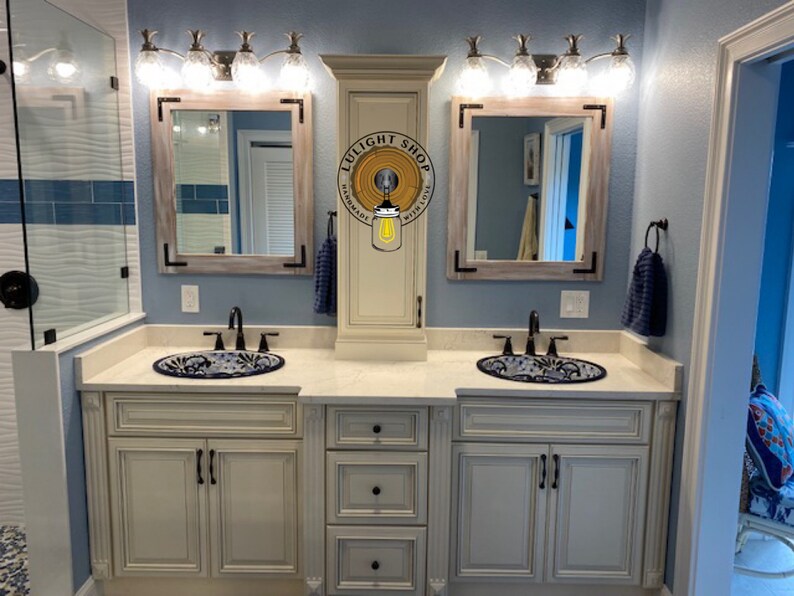 Pu&ograve; includere: Un mobile da bagno con due lavandini, specchi e lampade. Il mobile ha un mobile color crema con ferramenta nera. I lavandini hanno un motivo floreale blu e bianco. Gli specchi hanno una cornice in legno chiaro con accenti angolari neri. Le lampade hanno paralumi in vetro trasparente.