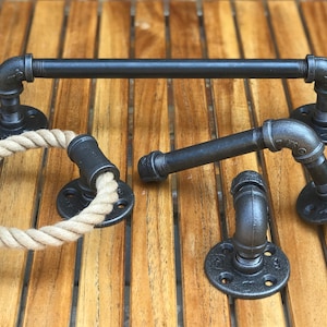 May include: A set of three black metal bathroom accessories, including a towel bar, a toilet paper holder, and a rope ring towel holder. The accessories are made of industrial pipe and have a rustic look.