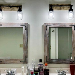Pu&ograve; includere: Due specchi da bagno rustici con cornici in legno e angoli in metallo. Sopra ogni specchio c'&egrave; un apparecchio di illuminazione con due paralumi a forma di barattolo di vetro trasparente. Gli apparecchi sono montati su una tavola di legno.