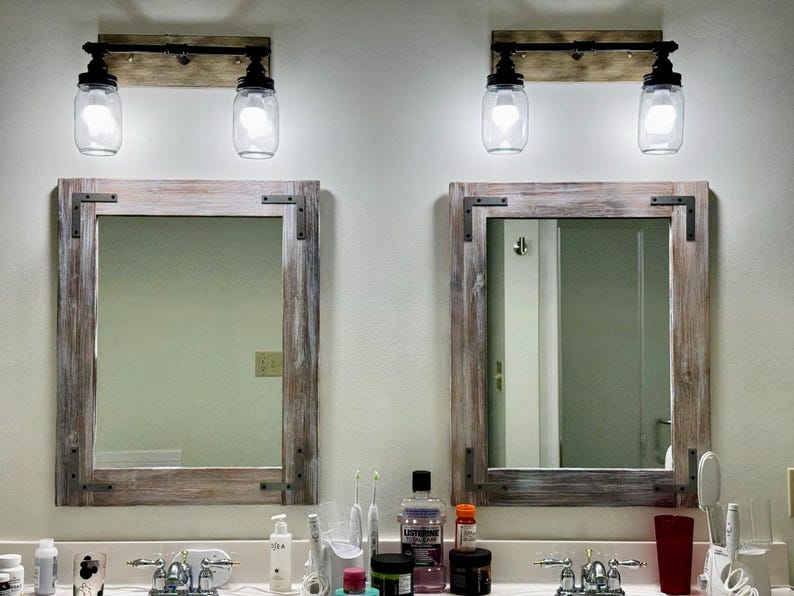Pu&ograve; includere: Due specchi da bagno rustici con cornici in legno e angoli in metallo. Sopra ogni specchio c'&egrave; un apparecchio di illuminazione con due paralumi a forma di barattolo di vetro trasparente. Gli apparecchi sono montati su una tavola di legno.