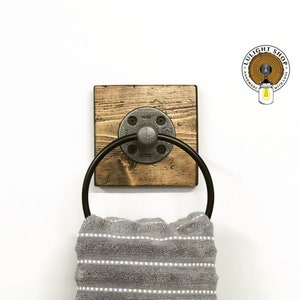 May include: A rustic wooden towel ring with a black metal ring and a pipe fitting. The ring is mounted on a white wall.