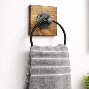 May include: A rustic wooden towel ring with a black metal ring and a pipe fitting. The ring is mounted on a white wall.