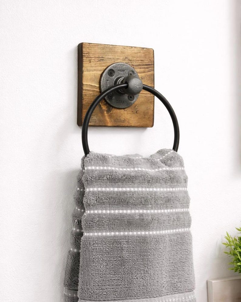 May include: A rustic wooden towel ring with a black metal ring and a pipe fitting. The ring is mounted on a white wall.